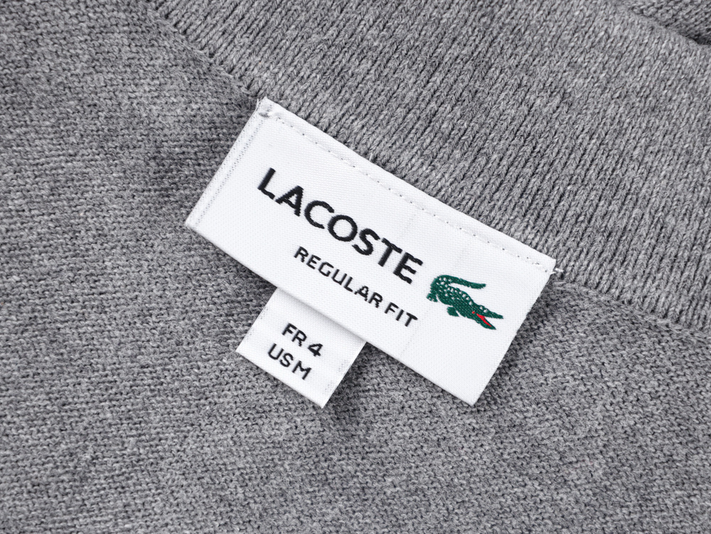 Gray knit fabric with a white rectangular label featuring black text and a green crocodile logo angled slightly upward.