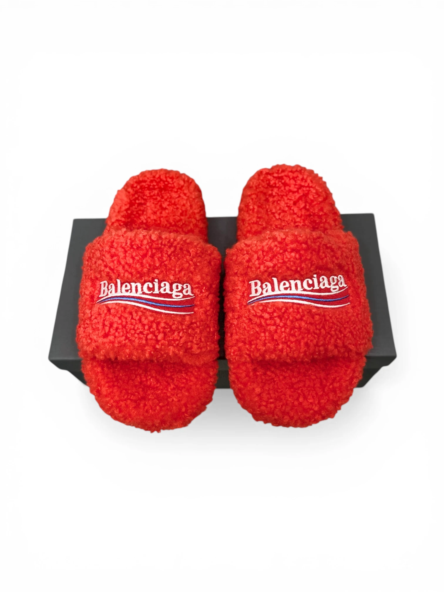 Red fluffy slippers with logo on strap, displayed on a black box.