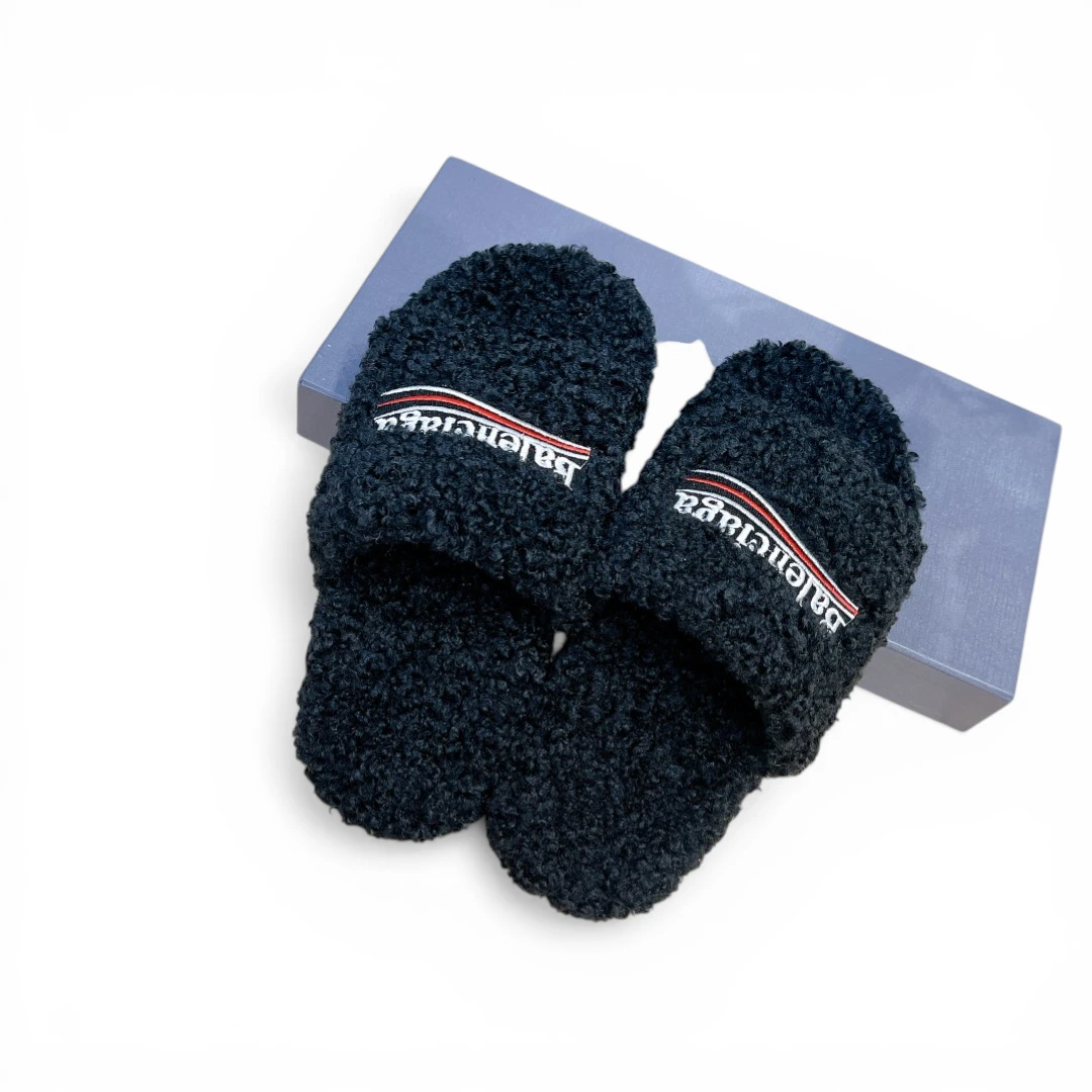 Black fuzzy slippers with embroidered logo, placed on a blue box.