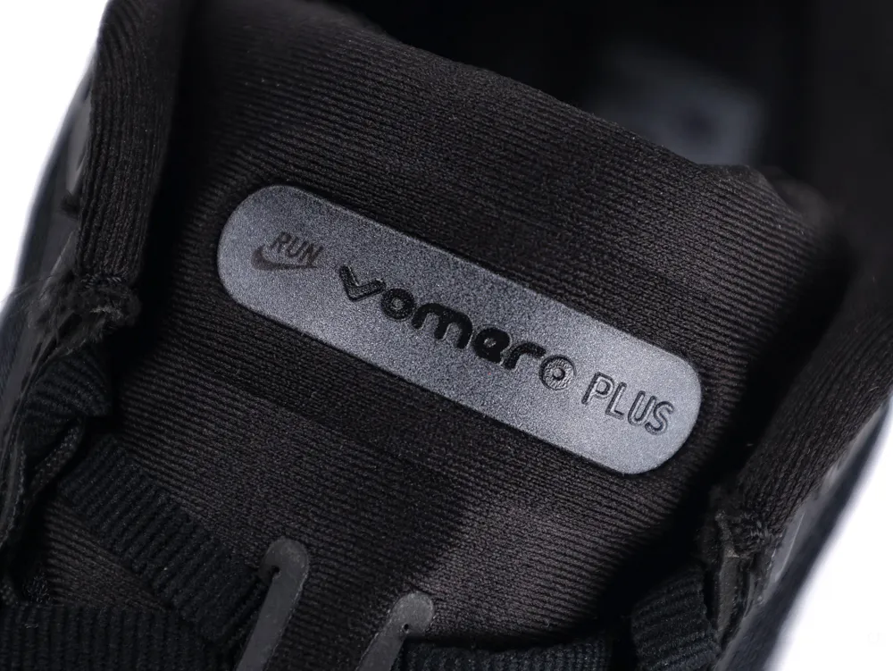 Black fabric sneaker tongue with gray plastic label featuring the text "Vomero Plus".